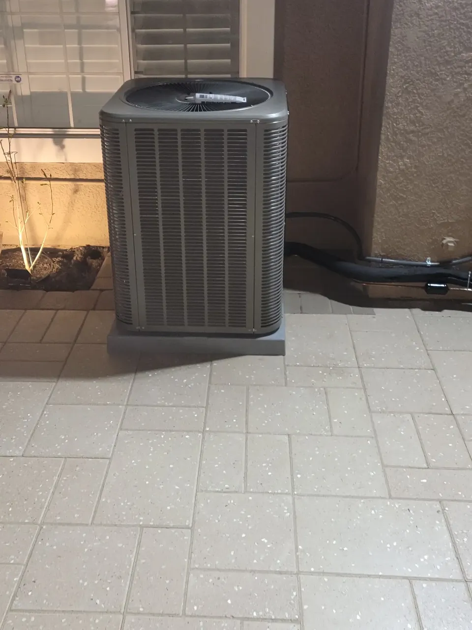 The outdoor unit of a split system standing on a patio beside a window with shutters