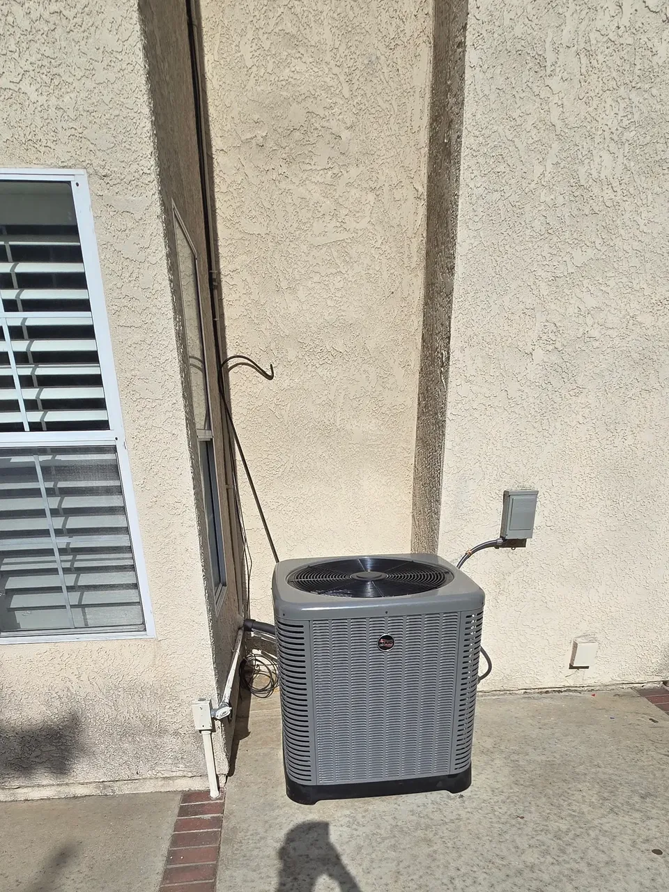 The outdoor unit of a mini-split standing against a house wall beside a shuttered window