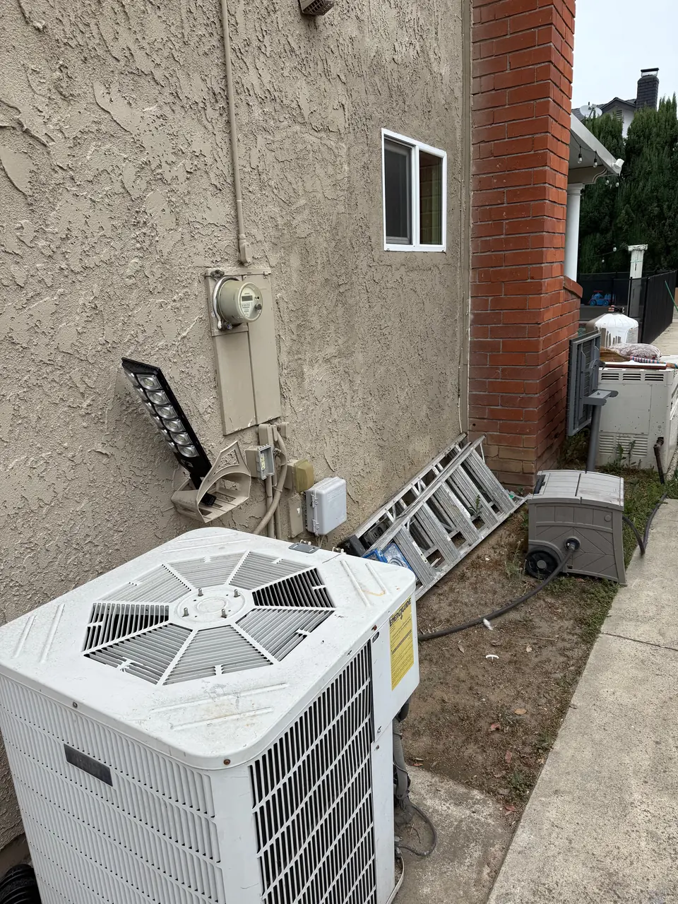 The outdoor unit of an air conditioner beside a house wall, with a ladder and an electrical meter behind it